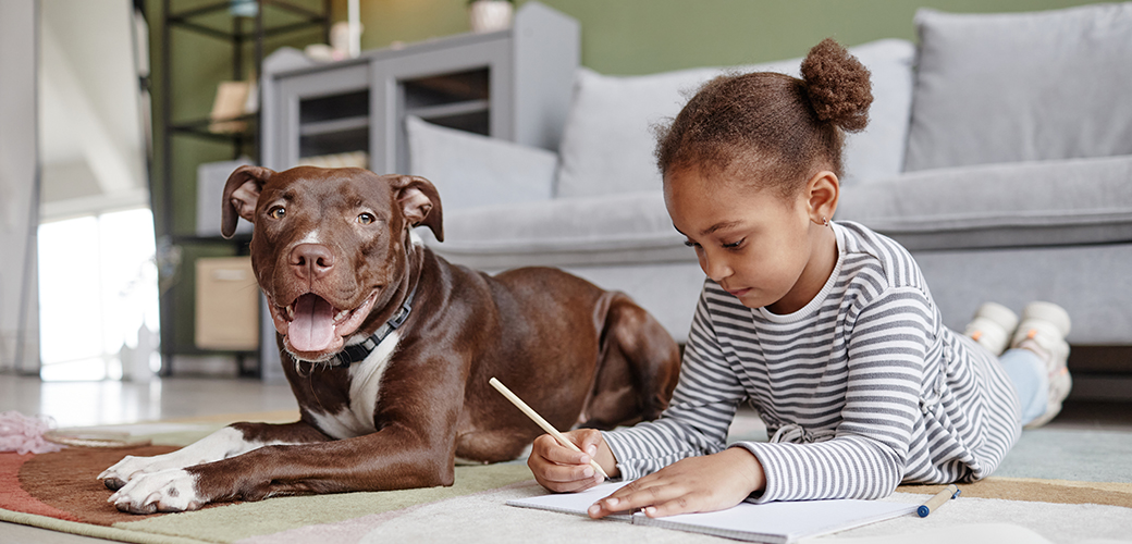 A little girl in a stripe shirt writing in a notebook on the floor next to large brown dog