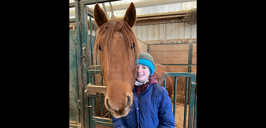 Felipe, a brown horse, and Clara in a blue winter coat and light blue and olive green hat in a stable together